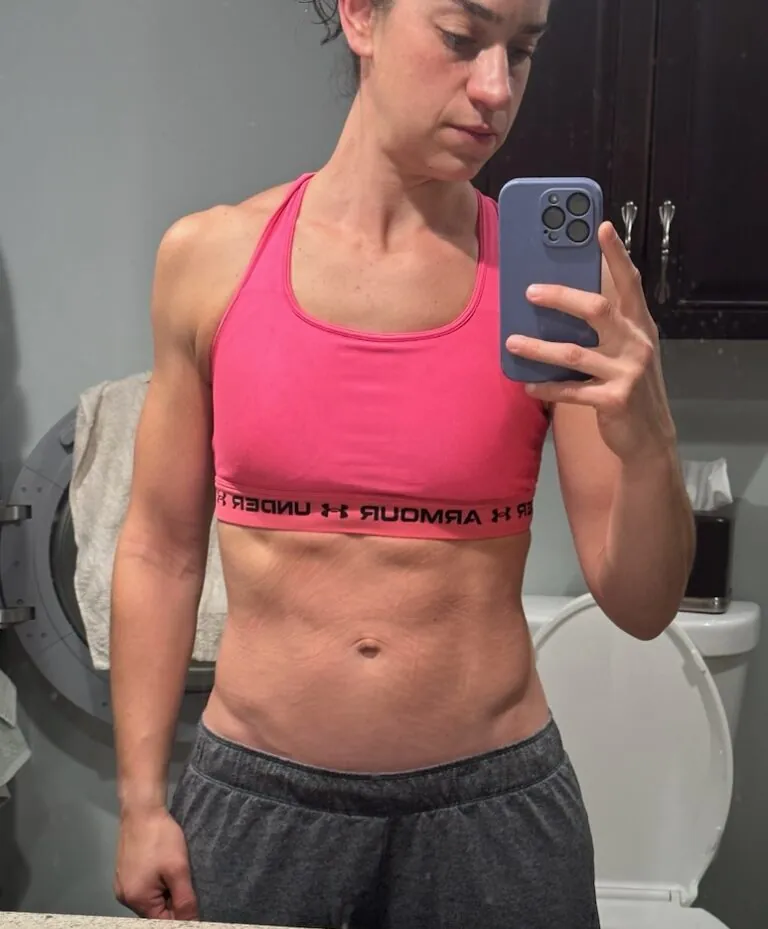 Person taking a mirror selfie in a bathroom, wearing a pink sports bra and gray sweatpants, showing a fit and muscular physique—proof that dedication to nutrition and workout routines pays off.
