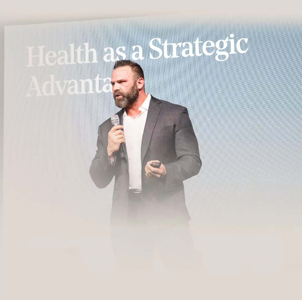 A man in a suit holds a microphone and presents on stage with the text "Health as a Strategic Advantage" displayed behind him, emphasizing the importance of nutrition and training for success.