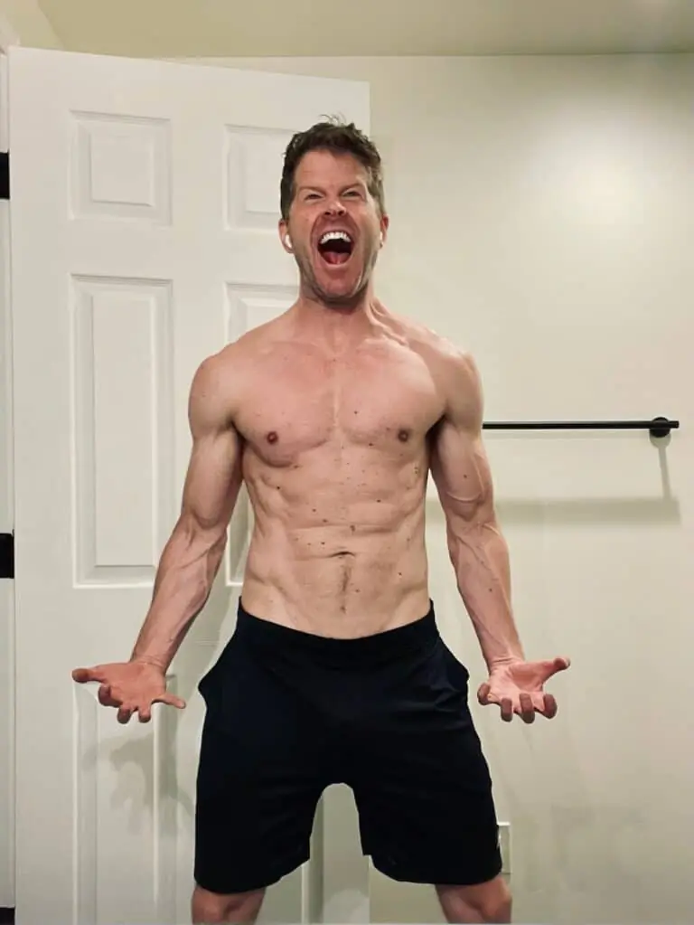 Shirtless man with defined muscles stands in front of a door, flexing his arms and shouting with his mouth wide open, showing off the results of intense fitness training.