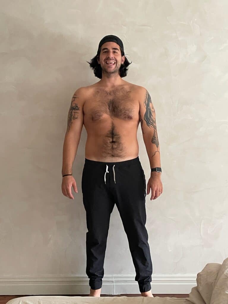 A man with long hair, wearing a backward cap and black shorts, stands shirtless against a plain wall, showing tattoos on both arms—he looks like a dedicated personal trainer ready for his next workout.