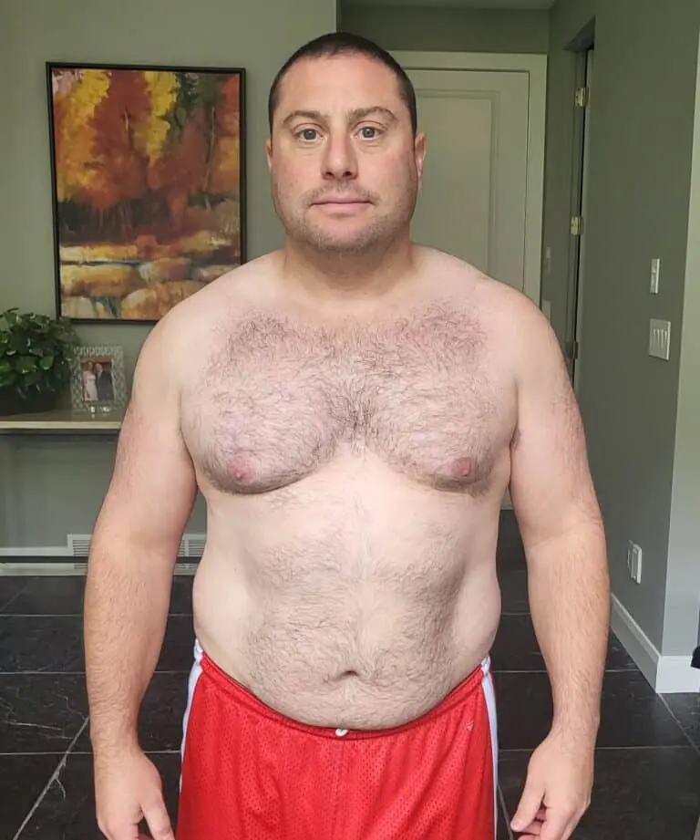 A man with short hair stands indoors wearing red shorts, facing the camera with a neutral expression. Behind him, a framed picture and plant rest on a table—an inviting scene that could suit an online fitness coach or nutrition expert.