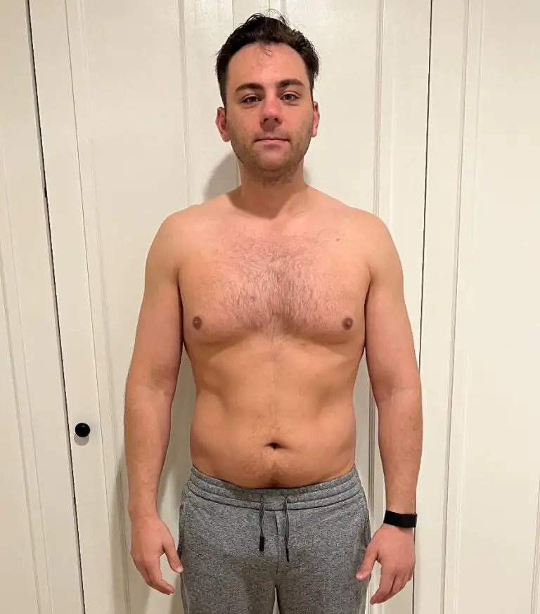 A man stands facing forward against a white paneled wall, shirtless and wearing gray sweatpants and a black wristband, with a neutral expression—resembling an online fitness coach or personal trainer ready to motivate.