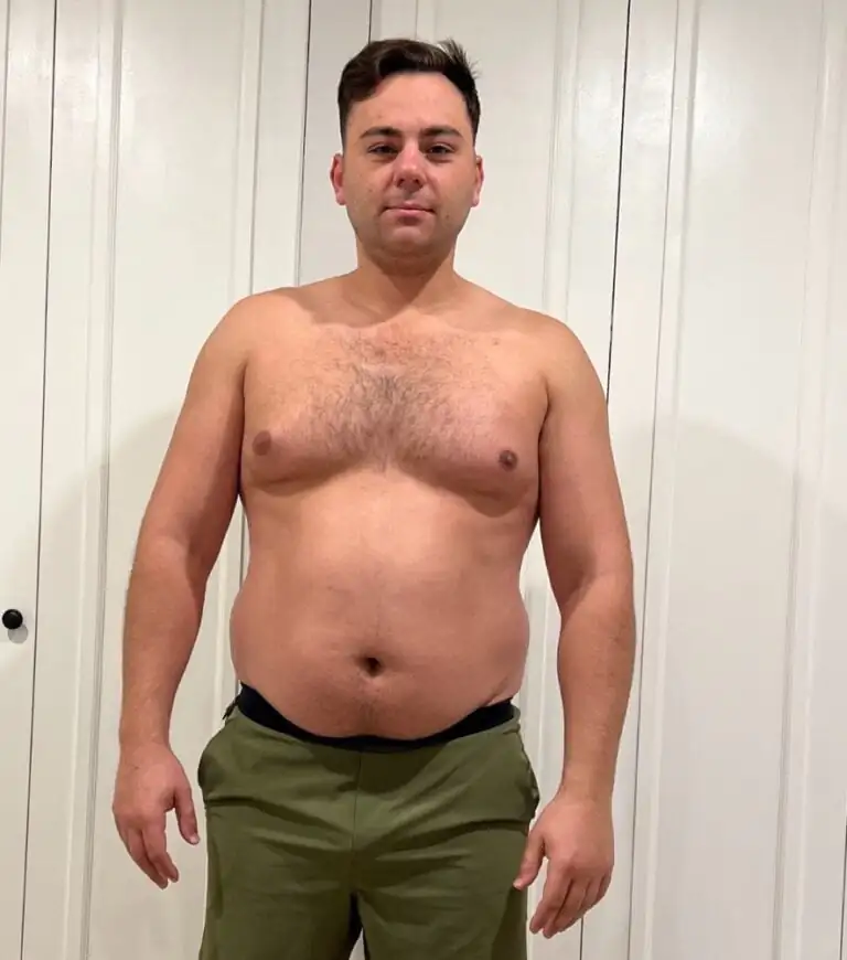 A man with short brown hair stands shirtless, wearing olive green pants, in front of white paneled doors—ready for his workout, he embodies the focus of an online fitness coach.