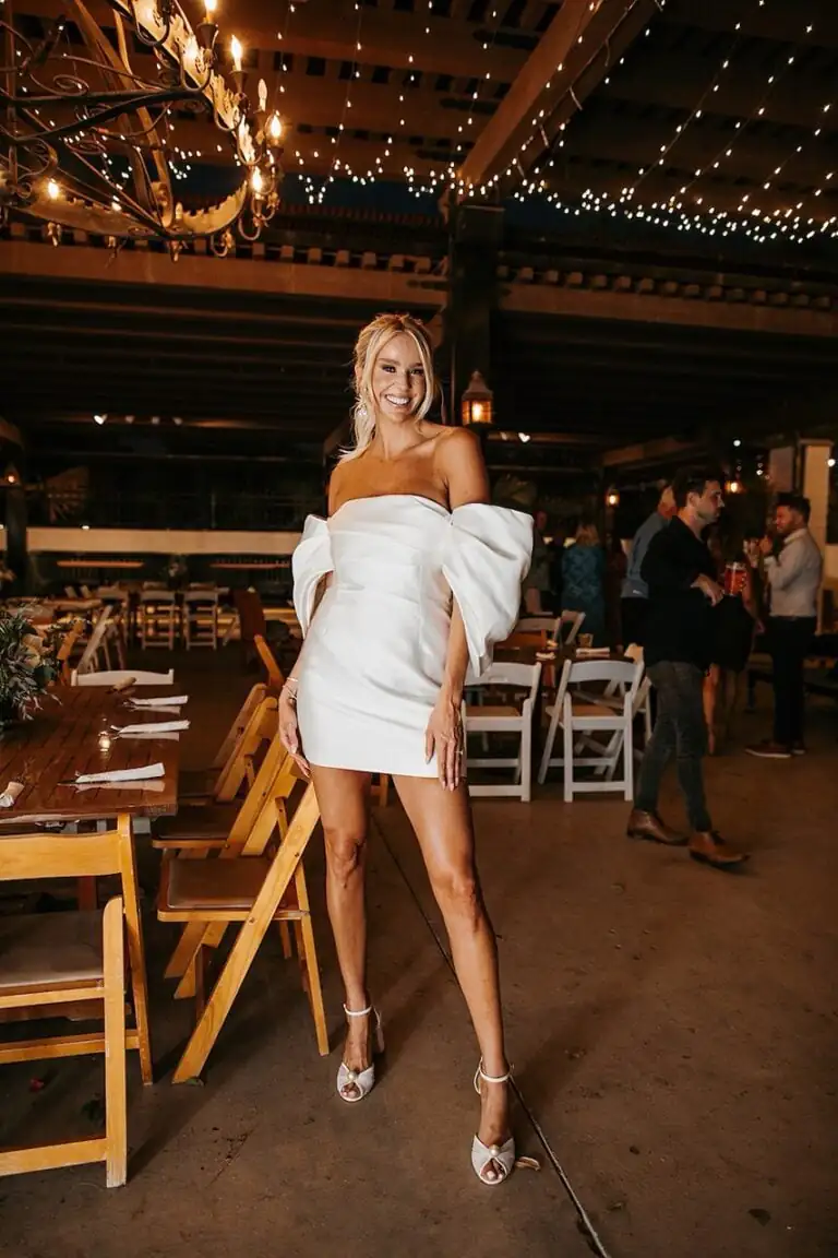 A woman wearing a white off-the-shoulder mini dress with large puff sleeves stands and smiles in a decorated indoor event space with wooden chairs and tables, fresh from her latest fitness training session.