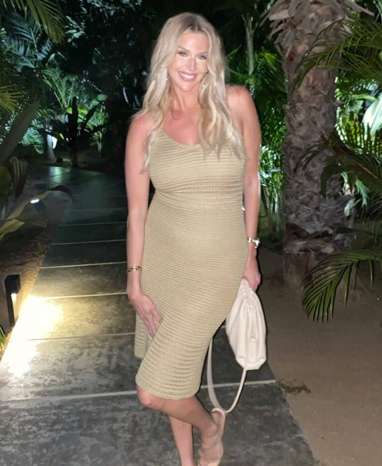 A woman in a beige sleeveless dress stands on an outdoor path at night, surrounded by tropical plants, smiling at the camera after a workout with her online fitness coach.