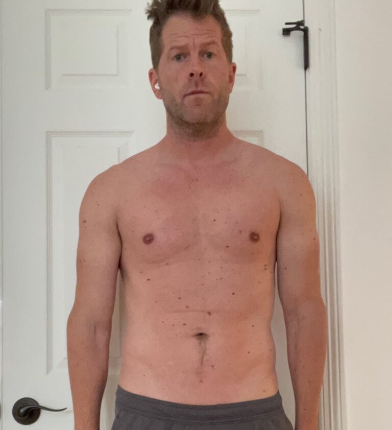 A shirtless man, possibly a personal trainer, stands indoors in front of a white door, looking directly at the camera with a neutral expression.