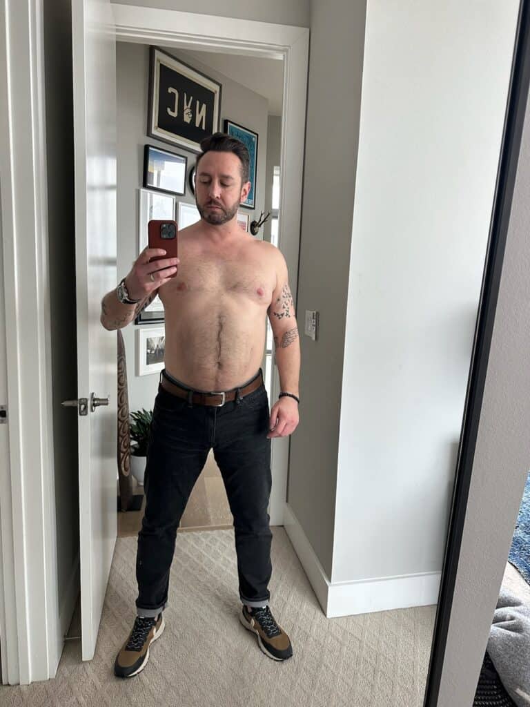 A shirtless man with short hair and tattoos on both arms takes a mirror selfie in a room decorated with framed pictures, showcasing his dedication to workout routines.