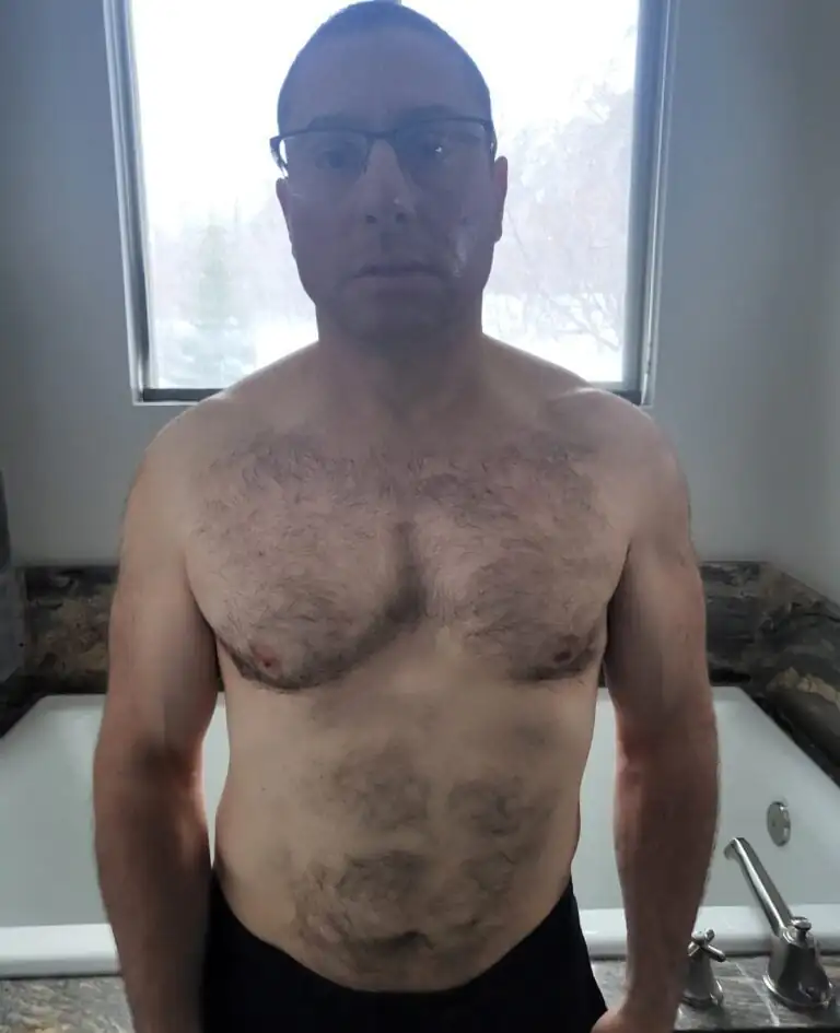 A man with short hair and glasses stands shirtless in a bathroom in front of a bathtub and window, looking directly at the camera—resembling an online fitness coach or personal trainer ready to share advice.