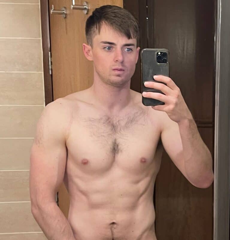 A shirtless man stands indoors, taking a selfie in a mirror with his phone, showing the results of his training and dedication to nutrition.