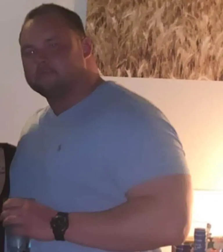 A man wearing a light blue t-shirt and black watch stands indoors, holding a drink, possibly after a workout, with a partial view of a person beside him.