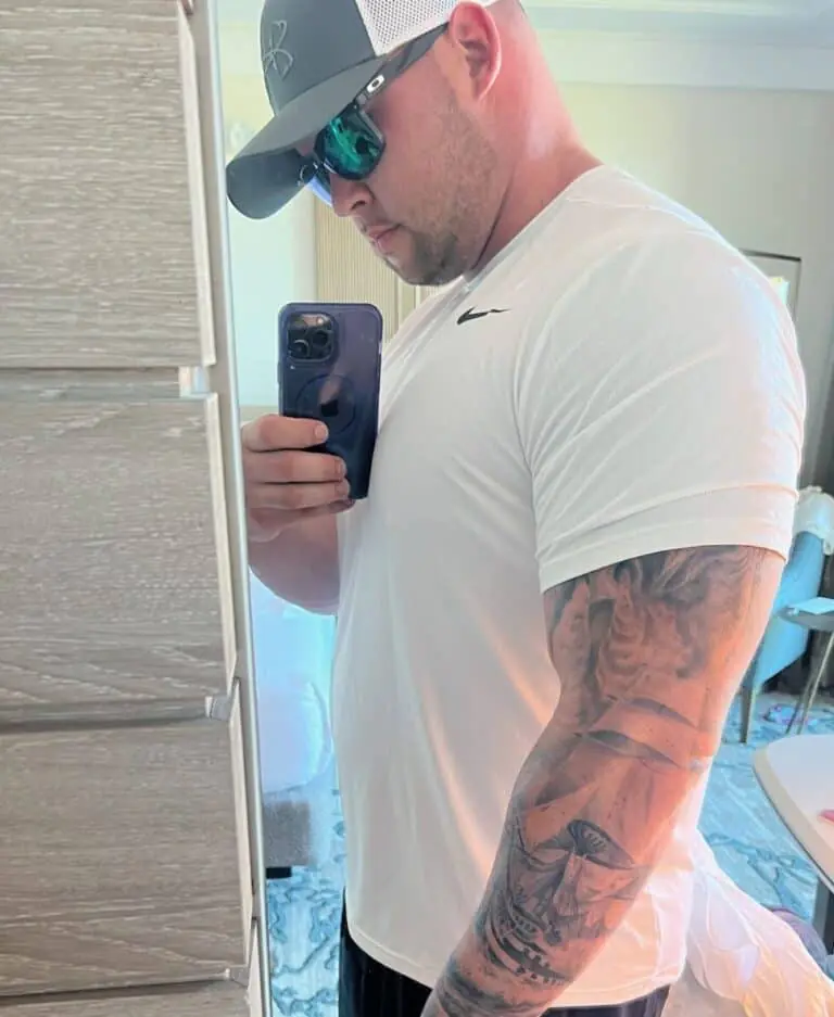 Man wearing sunglasses, a cap, and a white Nike t-shirt takes a mirror selfie, showing his tattooed arm and holding a phone—capturing the look of an online fitness coach.