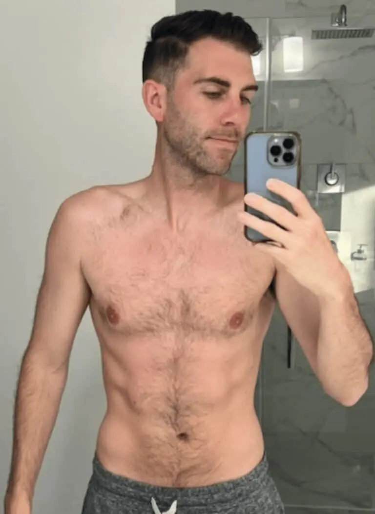 A man with short dark hair, possibly a personal trainer, takes a shirtless mirror selfie in a bathroom, wearing grey shorts and holding a smartphone, showcasing his fitness progress.