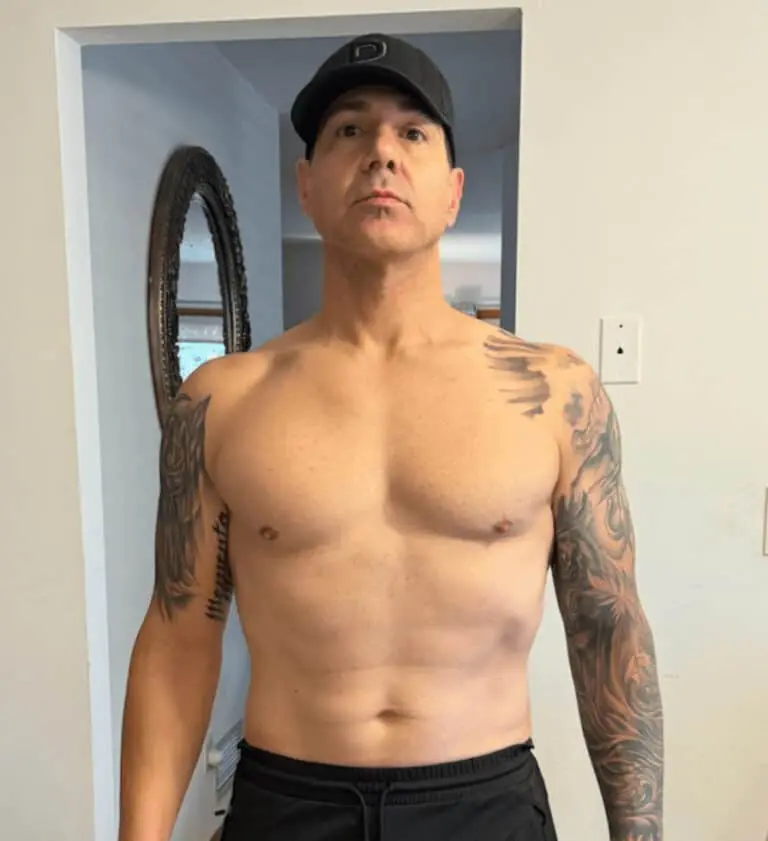 A man wearing a black cap and black shorts stands shirtless indoors, showing tattoos on both arms. A round wall mirror is visible in the background, suggesting he might be a personal trainer focused on fitness and nutrition.