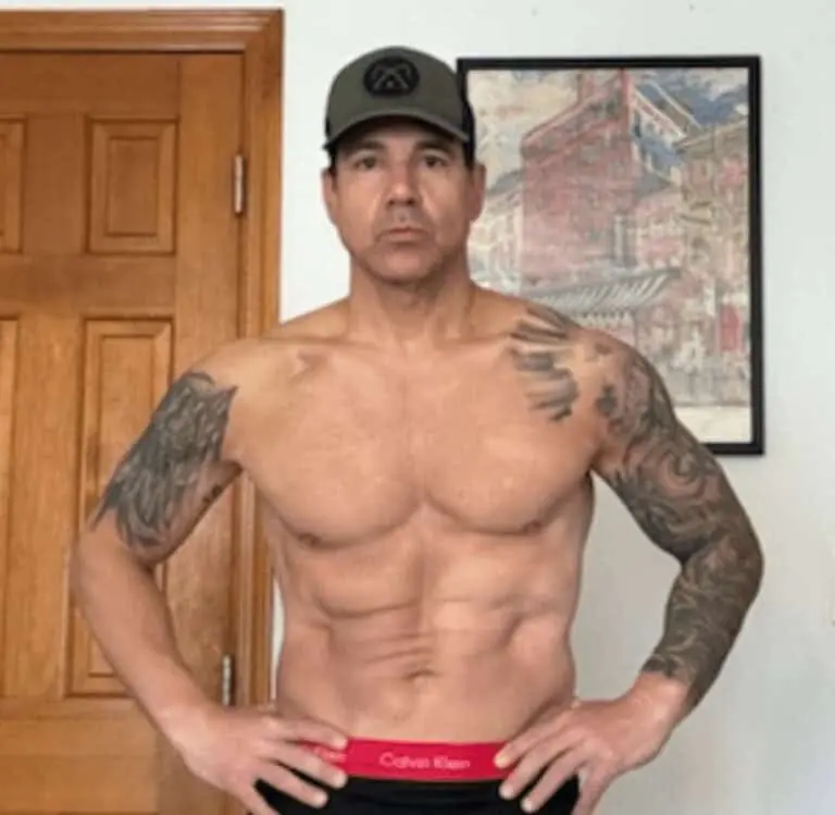 A shirtless man with tattoos poses indoors, showing off his fitness as he stands in Calvin Klein underwear and a cap in front of a wooden door and framed artwork.