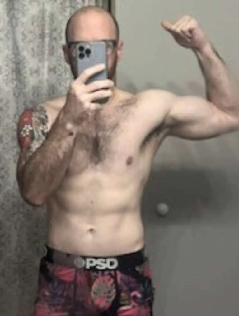 Shirtless person taking a mirror selfie, flexing one arm to show off their workout progress; wearing patterned shorts and holding a phone in front of their face.