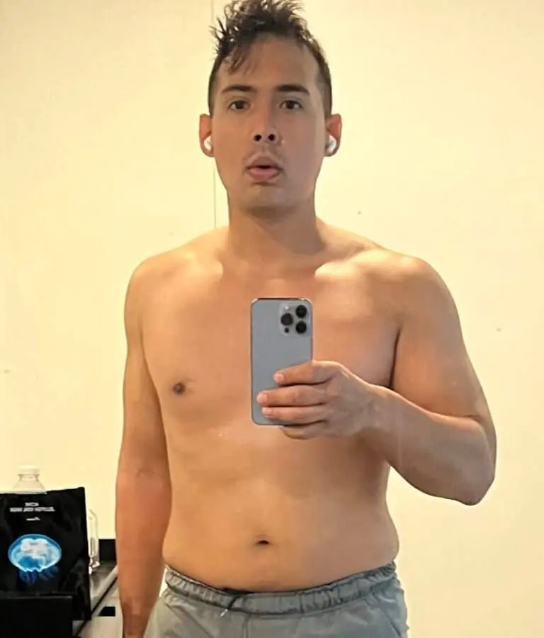 A shirtless person stands indoors, taking a mirror selfie with a smartphone. Wearing gray shorts and wireless earbuds, they capture their workout progress—perhaps for their online fitness coach or personal trainer.
