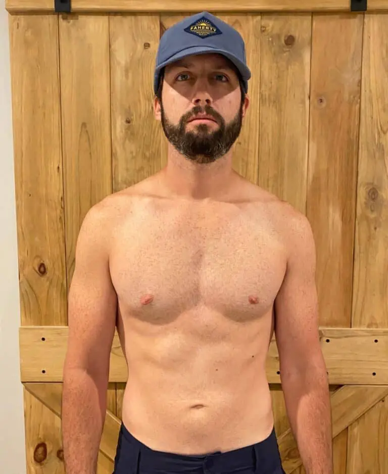 A bearded man wearing a blue cap stands shirtless in front of a wooden sliding barn door, facing the camera with a neutral expression—capturing the look of a dedicated personal trainer ready for his next workout.
