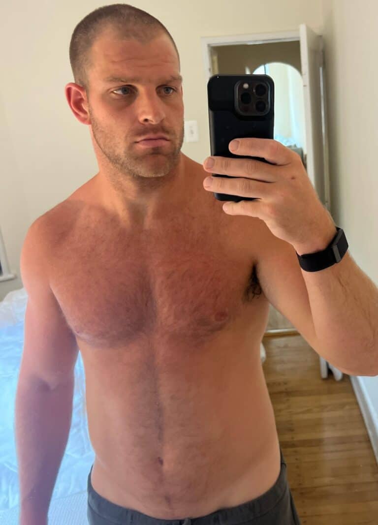 A shirtless man with short hair takes a mirror selfie in a room with light walls and wooden floors, proudly wearing a smartwatch—ready to track his training as an online fitness coach.