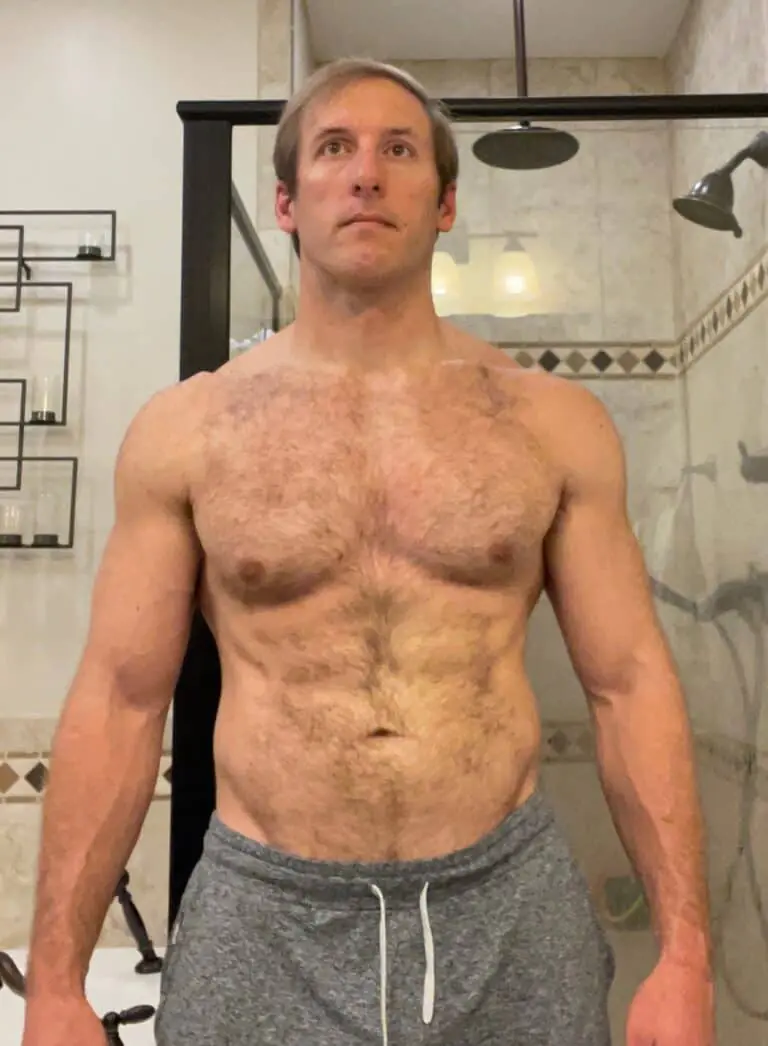A man with a muscular build stands shirtless in a bathroom, wearing gray drawstring shorts, showcasing his fitness. A glass shower and shelves are visible in the background.