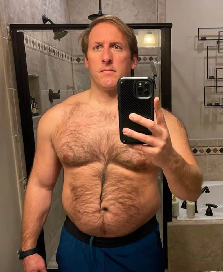 Man taking a mirror selfie in a bathroom, wearing blue shorts and holding a phone, with a neutral facial expression—capturing his progress from following an online fitness coach and dedicated workout routine.