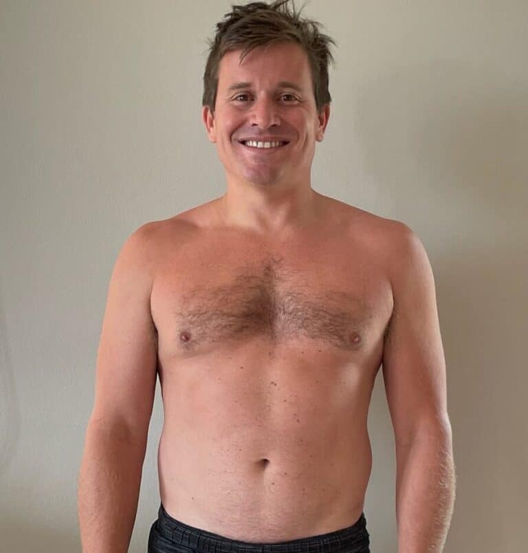 A smiling, shirtless man with short hair stands against a plain light-colored wall, wearing dark shorts—ready to start his training as an online fitness coach.