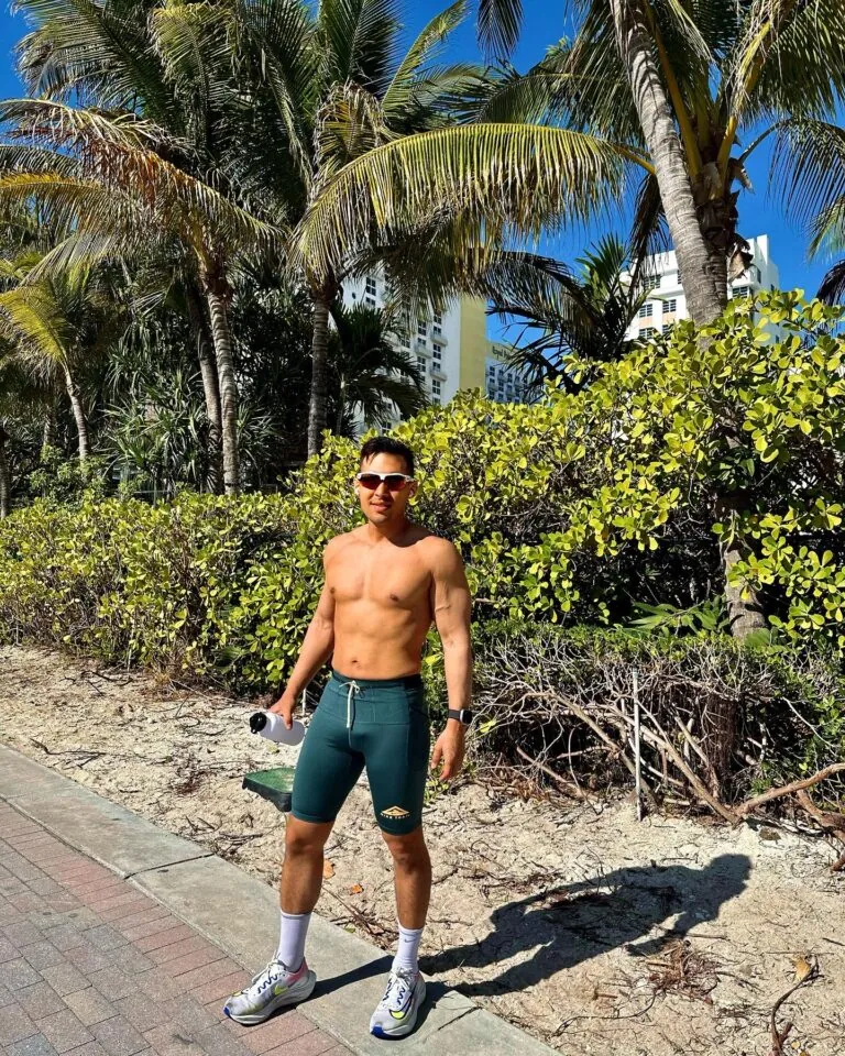 A man wearing sunglasses and swim trunks stands shirtless outdoors, holding a water bottle—ready for his workout—with green bushes and sunlight in the background.