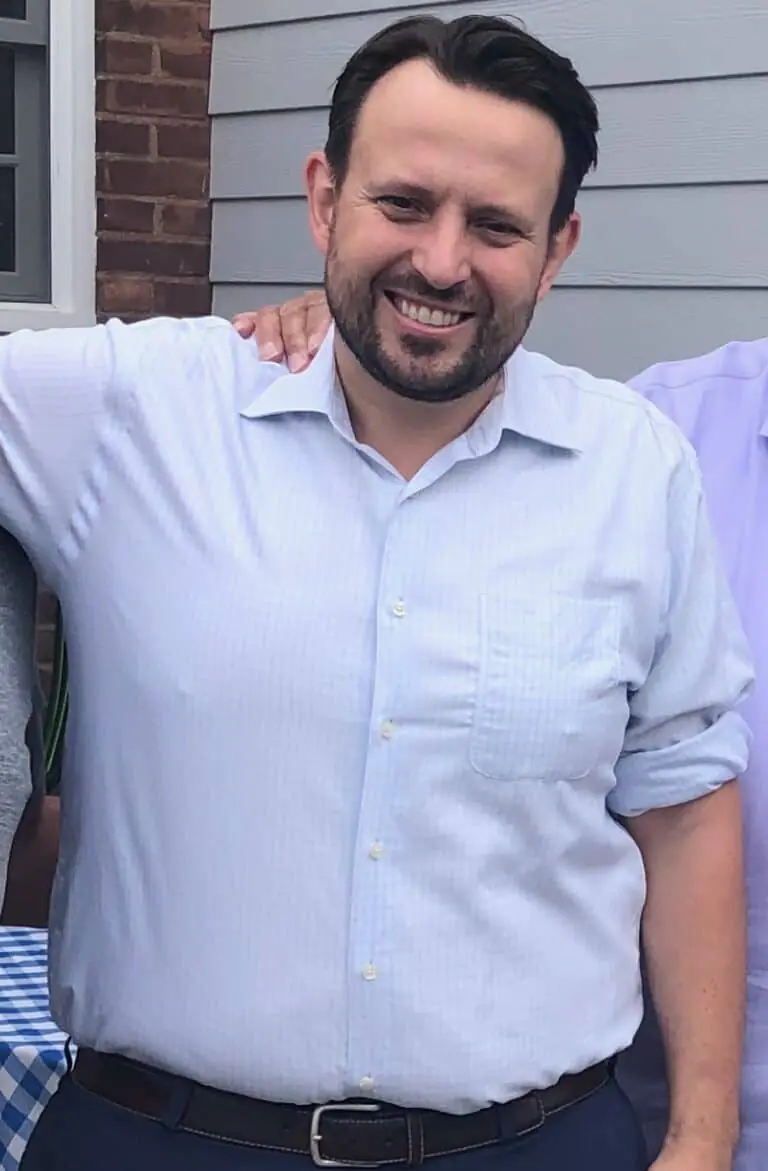 A man with dark hair and a beard, wearing a light blue button-down shirt and dark pants, stands outside in front of a gray siding wall, smiling at the camera—a personal trainer ready to inspire your fitness journey.