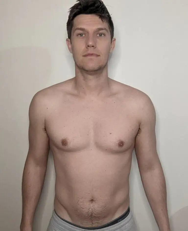 A shirtless adult man with short dark hair, possibly a personal trainer, stands facing forward against a plain white wall.