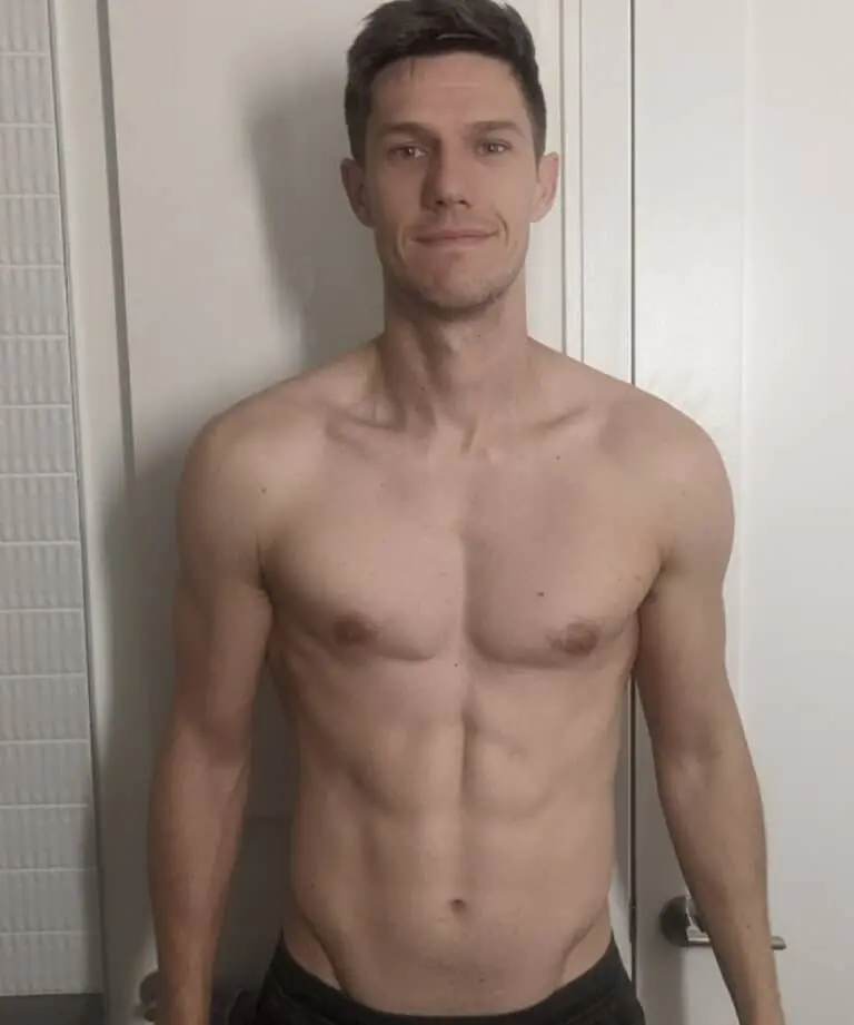 A shirtless man stands indoors in front of a white door, looking at the camera with a neutral expression, as if ready to start his workout or follow guidance from an online fitness coach.