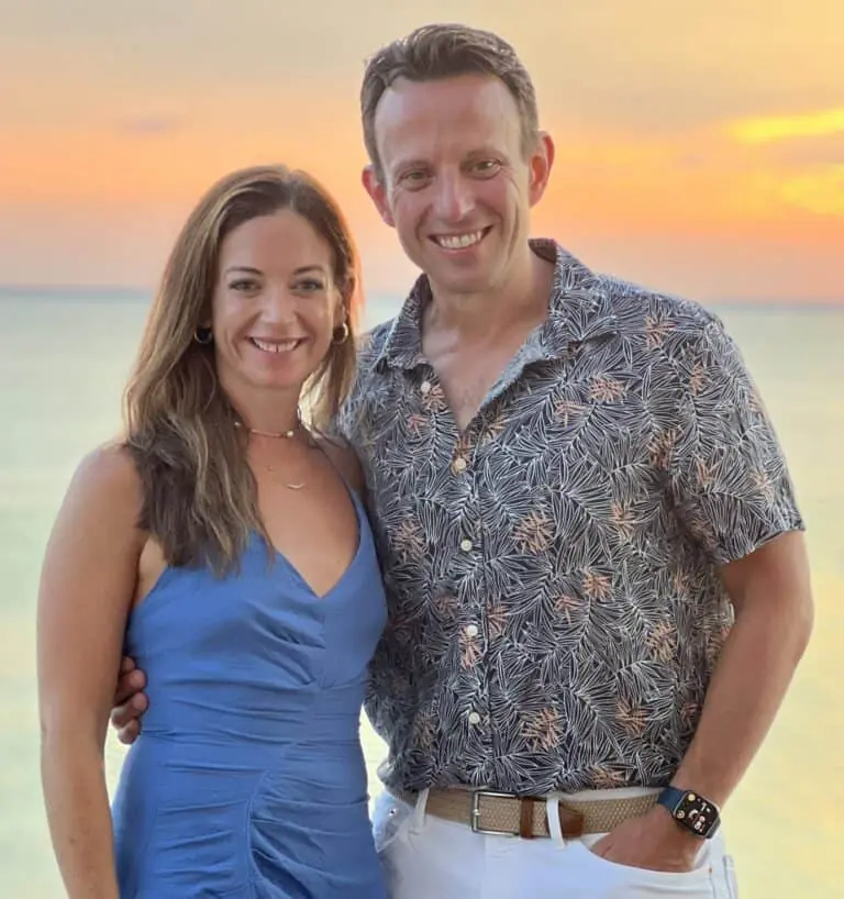 A man and woman stand together smiling in front of a sunset over the water, celebrating their fitness journey. The woman wears a blue dress and the man wears a patterned shirt and white pants.