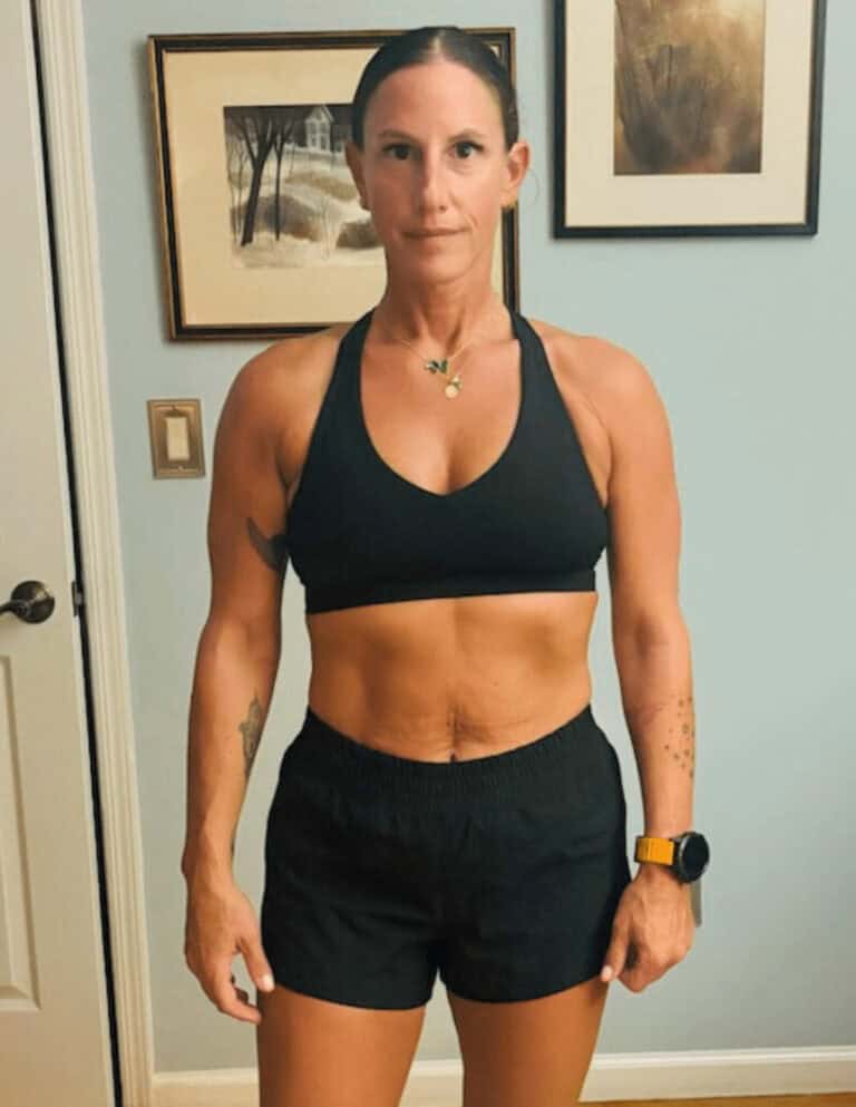 A woman in athletic wear stands indoors against a light blue wall with framed art, showing a fit and muscular physique—perfectly embodying the dedication of an online fitness coach.