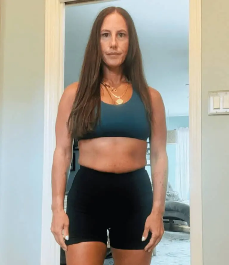 A woman with long brown hair stands indoors, showcasing her commitment to fitness in a blue sports bra and black shorts, facing the camera with a neutral expression.