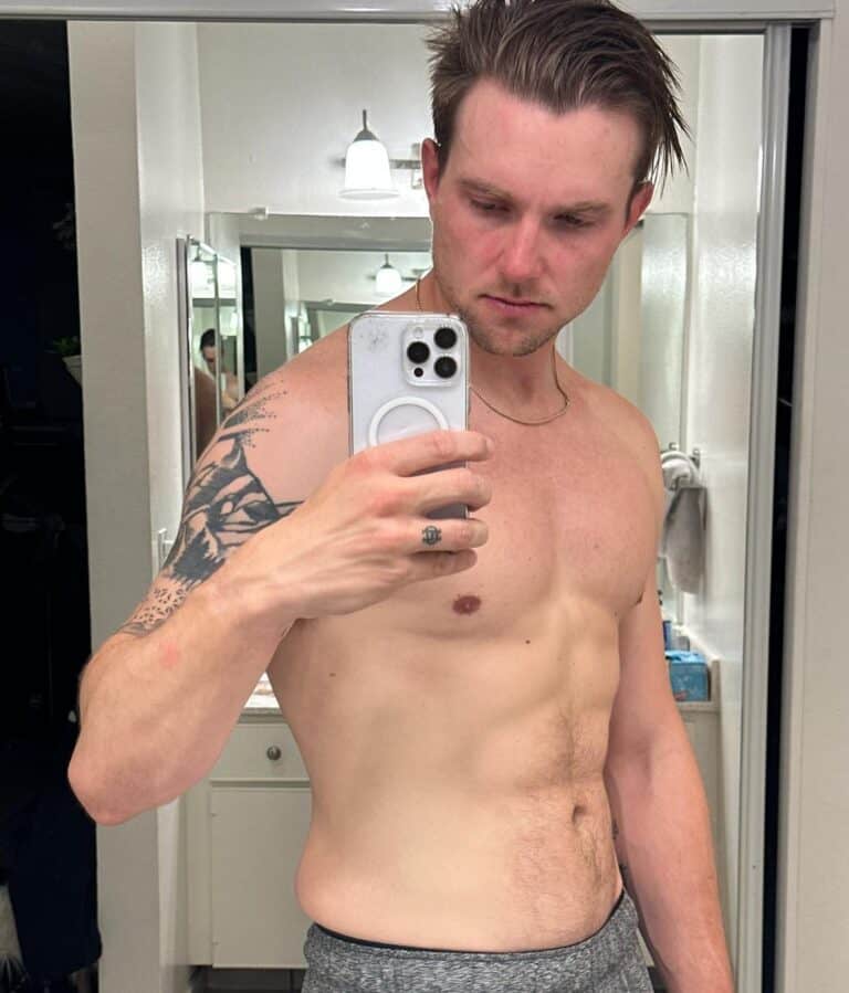 A shirtless person with a tattoo on their upper right arm, possibly a fitness enthusiast or personal trainer, takes a mirror selfie in a bathroom.
