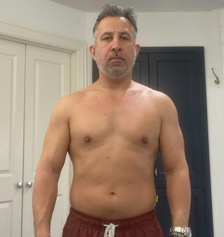 Middle-aged man with short gray hair and a beard standing shirtless indoors, wearing maroon shorts, facing the camera with a neutral expression—showcasing his fitness as a personal trainer.