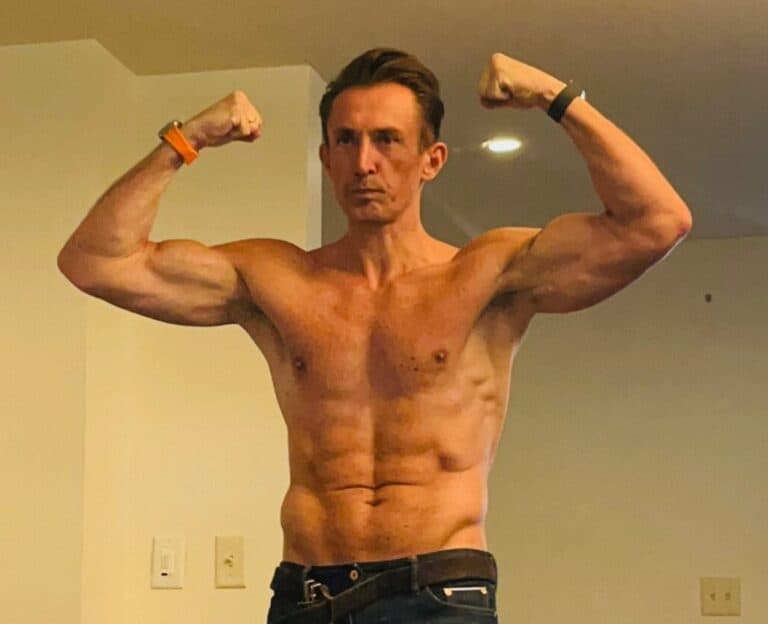 A shirtless man poses indoors, flexing his biceps with both arms raised—showcasing the results of dedicated workout training. He wears a watch and wristband in a well-lit room with beige walls.