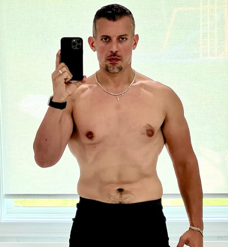 A shirtless man with short hair takes a post-workout selfie in front of a window, holding a smartphone in his left hand and wearing a necklace and wristwatch.