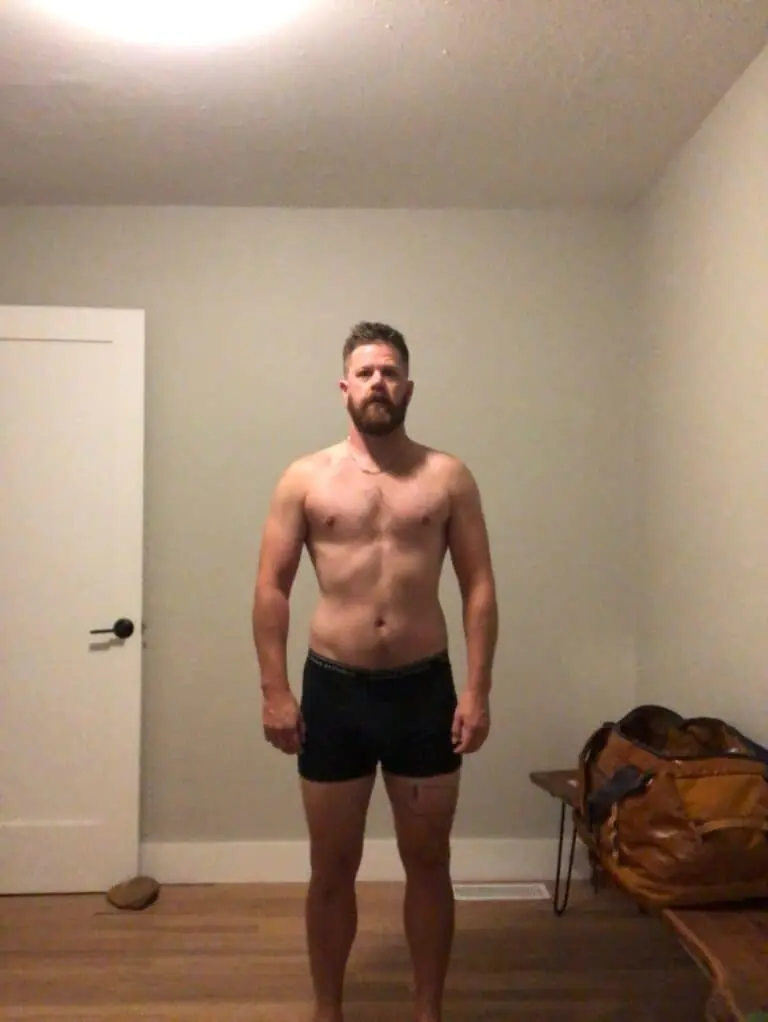 A bearded man with short hair, embodying fitness, stands facing forward against a plain wall, wearing black boxer briefs.