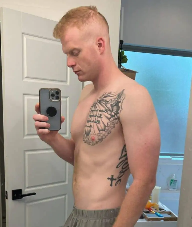 A shirtless person with short blond hair takes a mirror selfie in a bathroom, showing off training tattoos on their chest and side.