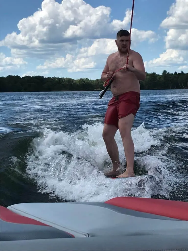 A man in red swim trunks holds a rope handle while standing on water, likely wakeboarding or water skiing, showcasing an intense workout with trees and clouds in the background.