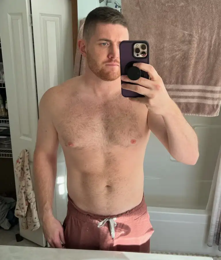 A fitness enthusiast with short hair and a beard takes a shirtless mirror selfie in a bathroom, wearing drawstring shorts and holding a phone with a pop socket.