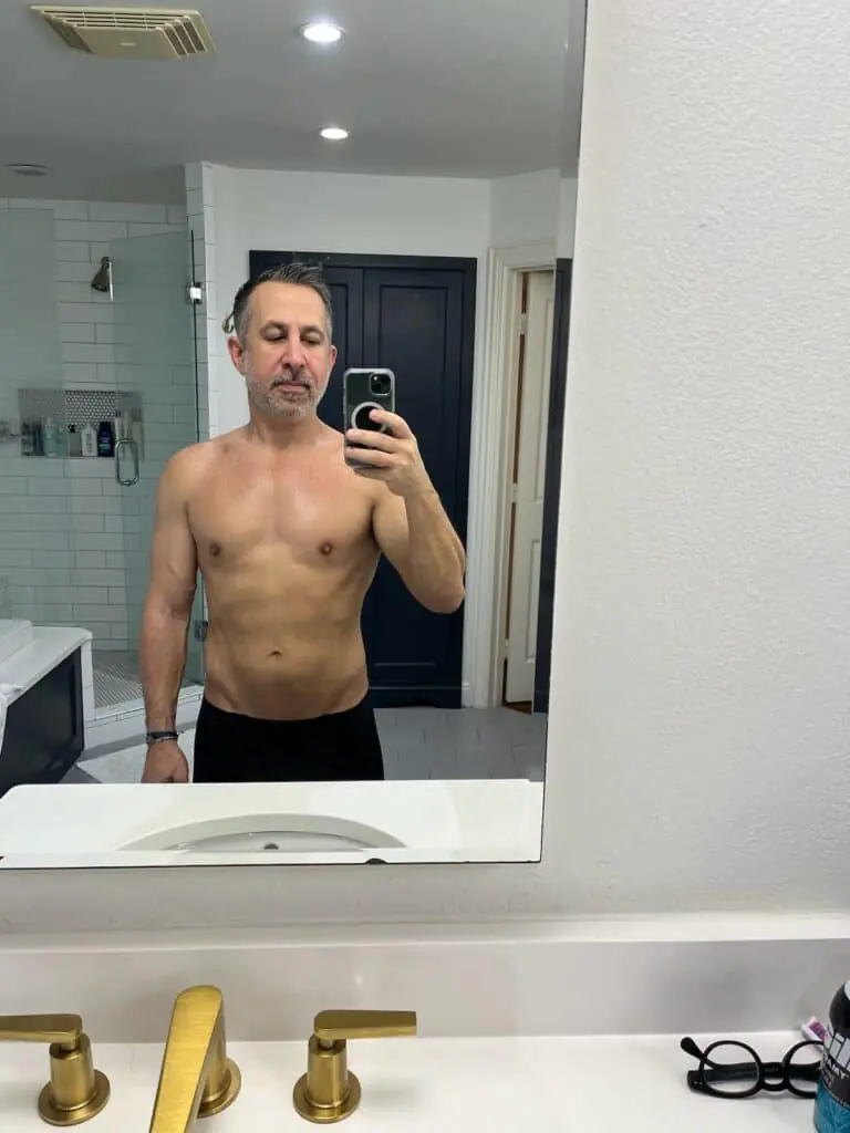 A middle-aged man with short gray hair and a beard takes a shirtless mirror selfie in a bathroom, showing off his fitness progress after a workout.