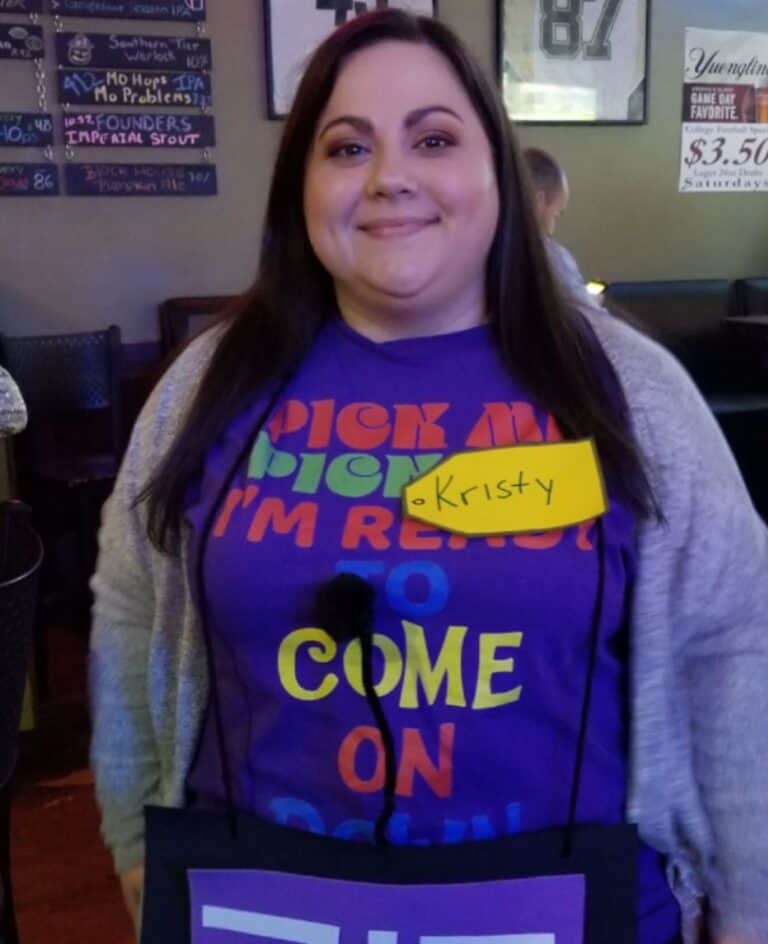 A woman with long dark hair, wearing a colorful “Pick Me I’m Ready To Come On Down” shirt and a yellow name tag labeled “Kristy,” stands in a bar or restaurant, ready for fun—and maybe some fitness tips.