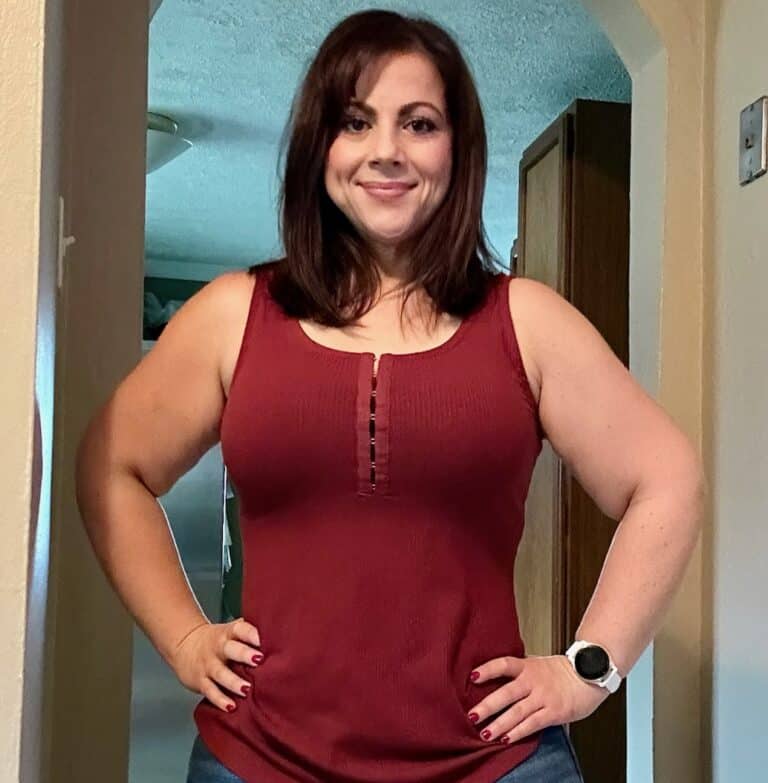 Woman with shoulder-length brown hair, wearing a sleeveless maroon top and a white smartwatch, stands indoors with her hands on her hips, looking at the camera—embodying confidence and fitness.