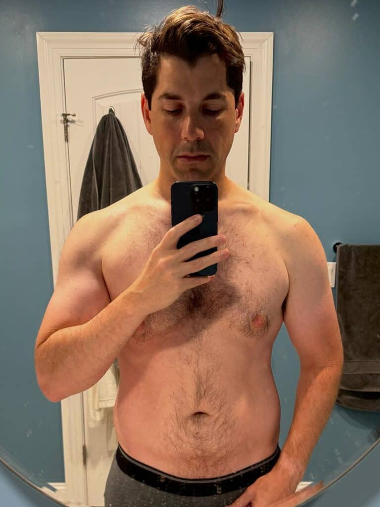 A man stands shirtless in front of a bathroom mirror, taking a selfie with his phone. Towels hang on racks behind him against a blue wall, showcasing the progress of his workout routine with guidance from an online fitness coach.
