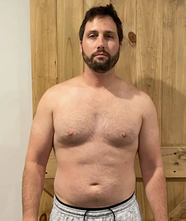 A man with short brown hair and a beard stands shirtless in front of a wooden door, wearing light gray shorts. As an online fitness coach, he looks straight at the camera with a neutral expression, ready to share workout tips.