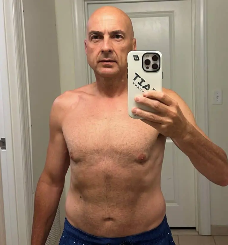 Middle-aged man takes a shirtless mirror selfie in a bathroom, holding a phone with a white case; white door and sink visible in the background, showing progress from his online fitness coach training.