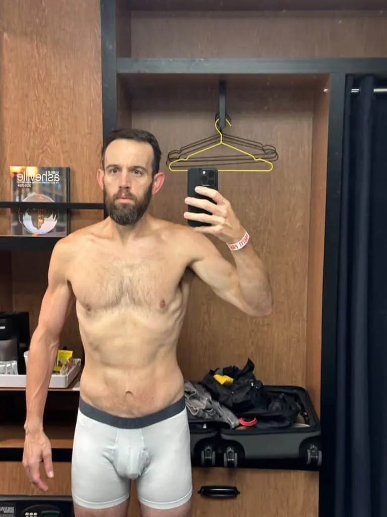 A bearded man stands shirtless in front of a mirror, taking a selfie after his workout in a room with wooden walls, clothing, and a book titled "Asheville" visible in the background.