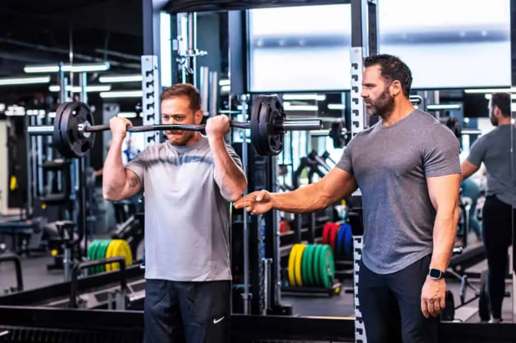 A man lifts a barbell in a gym while another man stands nearby, observing and offering guidance during the workout.