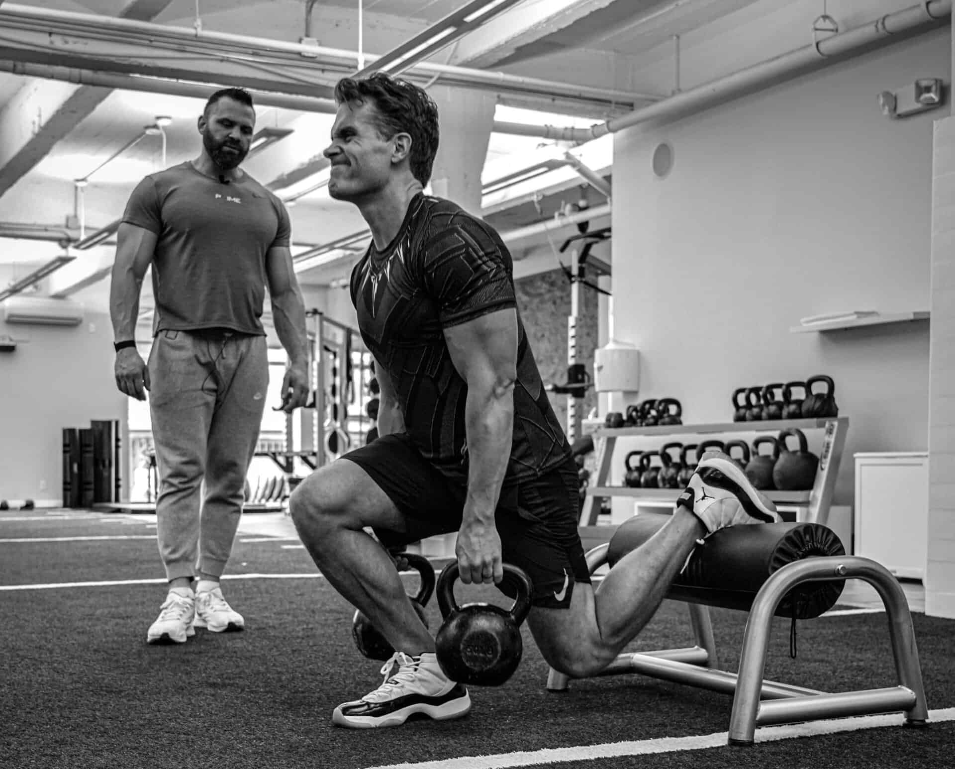 A man performs a Bulgarian split squat with kettlebells as another observes his workout in a gym setting.