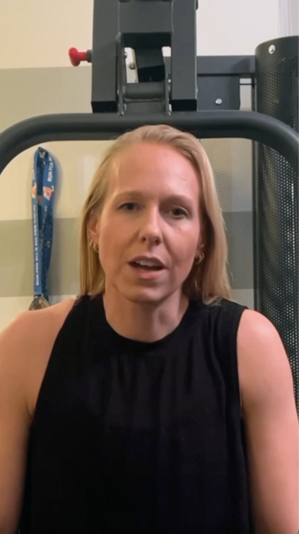 A woman with straight blonde hair, wearing a sleeveless black top, sits indoors with exercise equipment and a medal in the background—reflecting her dedication as an online fitness coach.